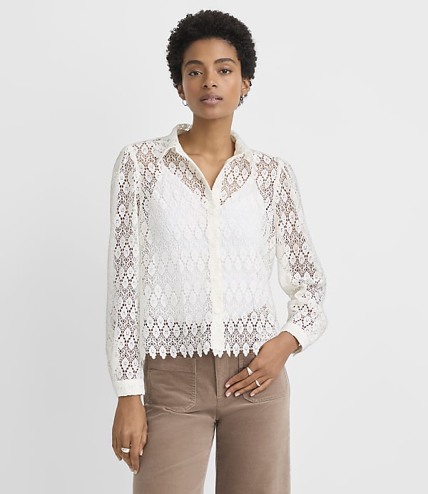 A woman in a white lace blouse and brown pants stands against a plain white background.