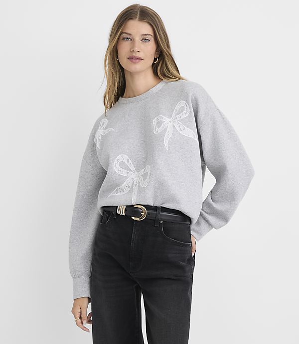 A woman in a grey sweatshirt with bow designs and black jeans stands against a plain grey background.