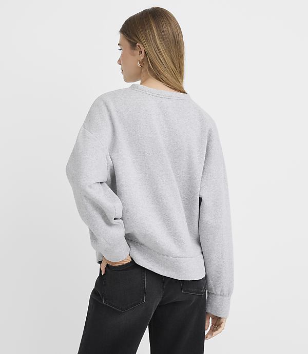 A woman in a grey sweatshirt and black jeans stands with her back to the camera against a plain white background.