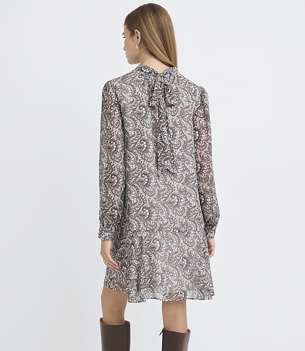A woman in a patterned dress stands against a plain background.