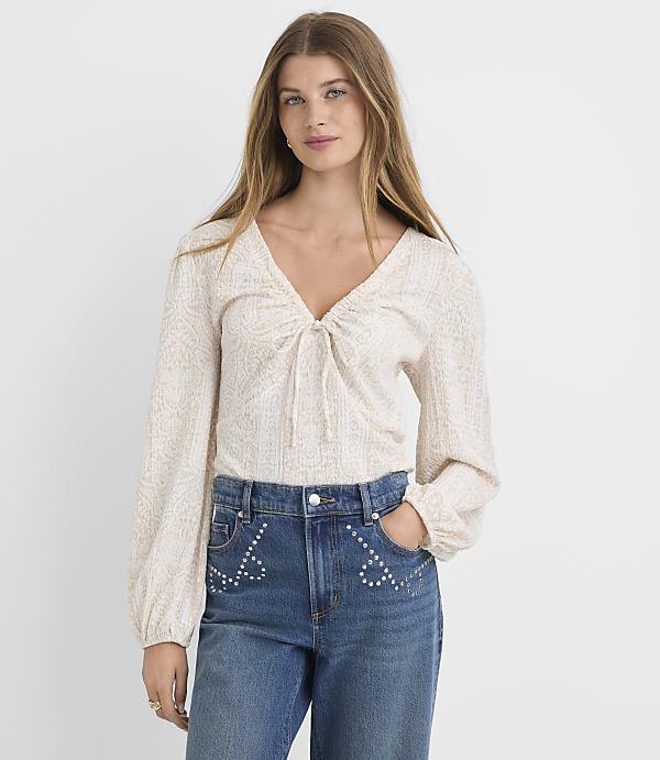 A woman in a patterned blouse and embellished jeans stands against a plain white background.