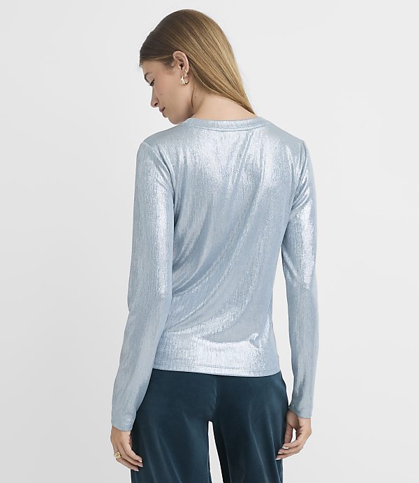 A woman in a shimmering blue long-sleeved top and teal velvet pants stands against a plain, light blue background.