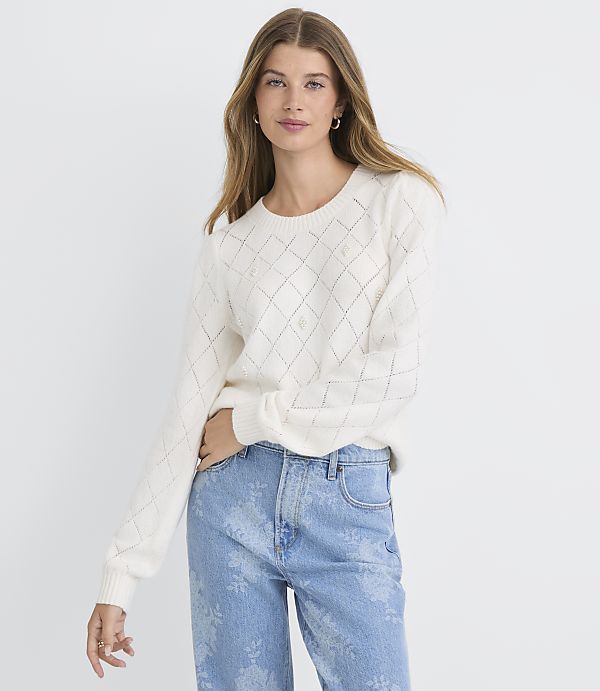 A woman in a cream-colored argyle sweater and light blue patterned jeans stands against a plain white background.