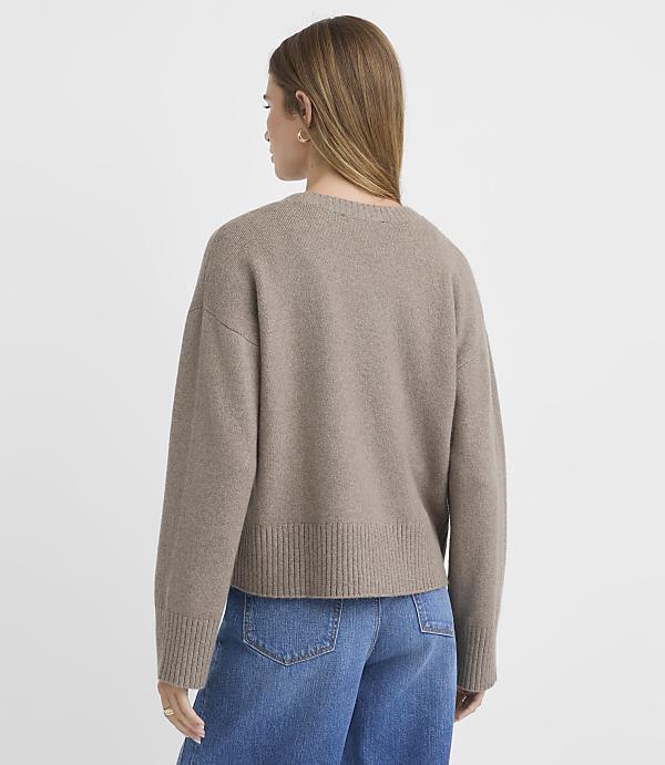 A woman in a beige sweater and blue jeans stands against a plain white background.