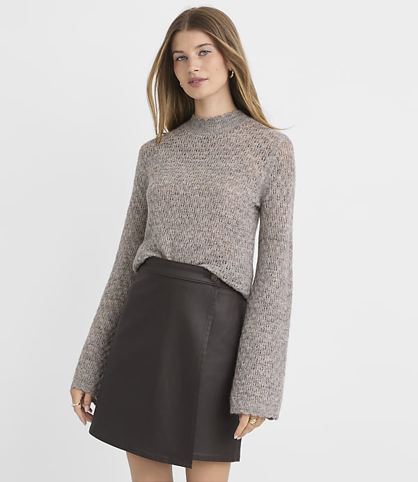 A woman in a grey knit sweater and a dark brown faux leather skirt stands against a plain white background.
