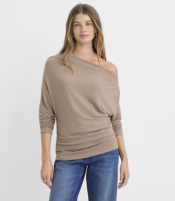 A woman in a taupe off-the-shoulder top and blue jeans stands against a plain white background.