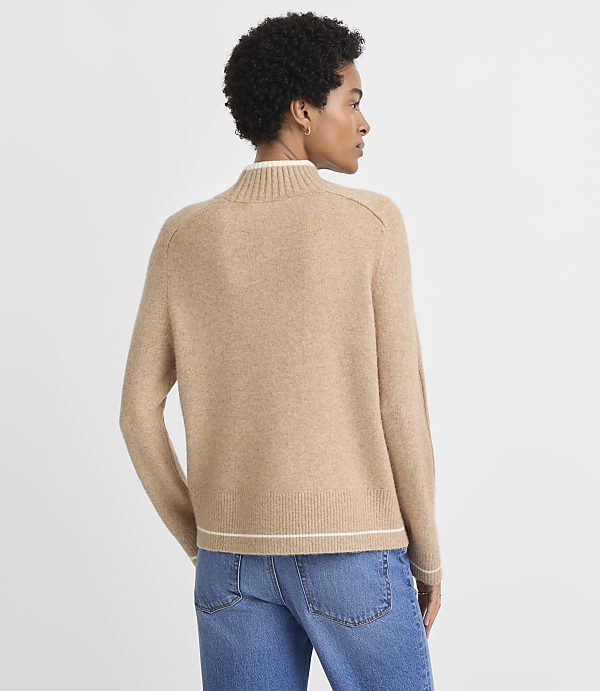 A woman in a tan mock-neck sweater and blue jeans stands against a plain, light gray background.
