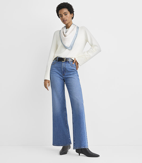 A woman in a white sweater, blue jeans, and black boots stands against a plain white background.
