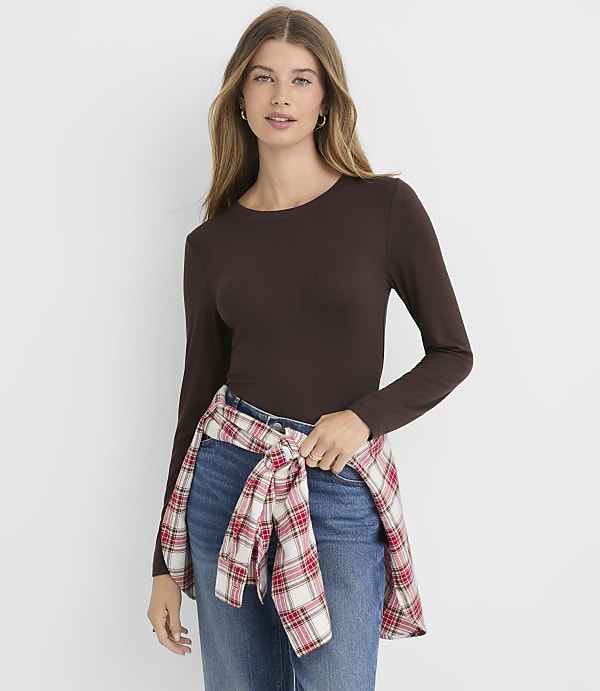 A woman in a brown long-sleeved shirt and jeans stands against a plain white background, with a red and white plaid shirt tied around her waist.