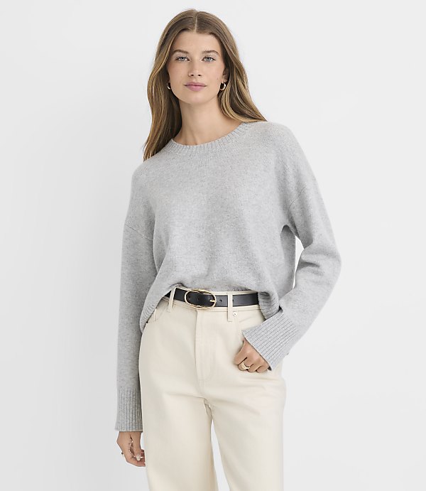 A woman in a light gray sweater and cream-colored pants stands against a plain white background.