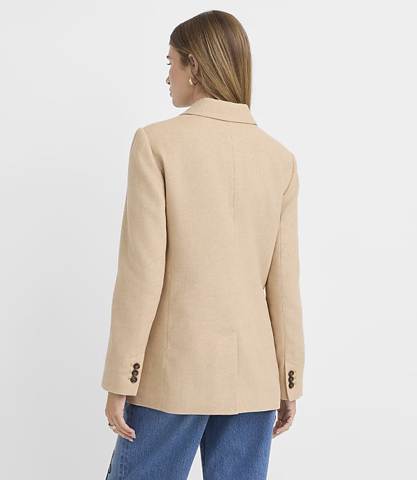 A woman in a tan blazer and blue jeans stands against a plain white background.