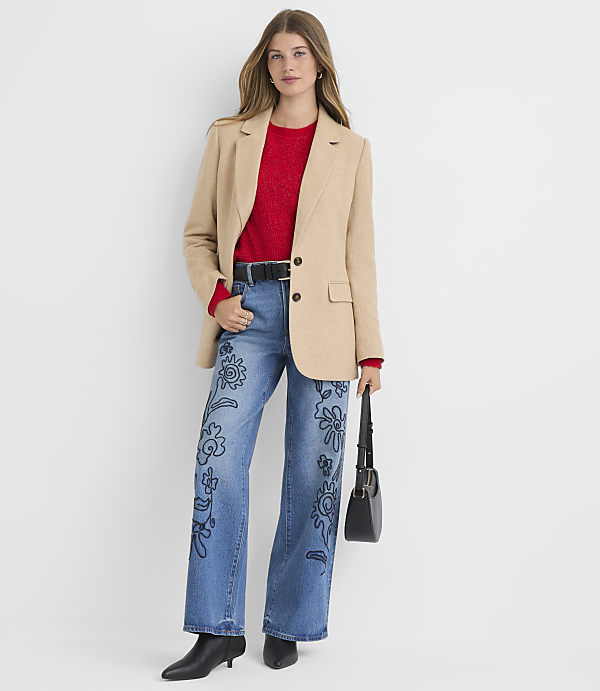A woman in a beige blazer, red sweater, and embroidered jeans stands against a plain white background, holding a black handbag.