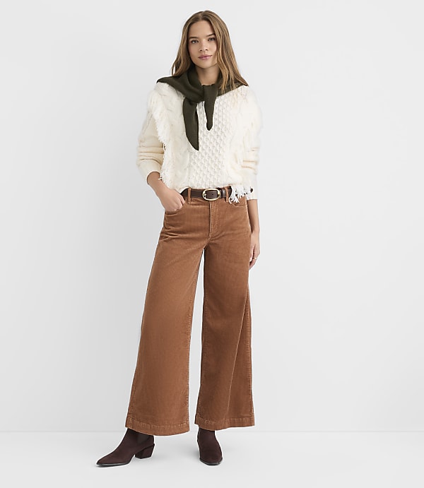 A woman in a cream cable-knit sweater, olive green scarf, and brown corduroy pants stands against a plain white background.