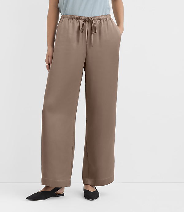 A person wearing loose, taupe, wide-leg pants with a drawstring waist and black pointed-toe flats stands against a plain white background.