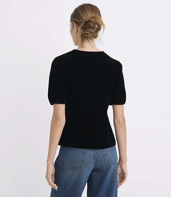 A woman in a black velvet top and blue jeans stands against a plain white background.