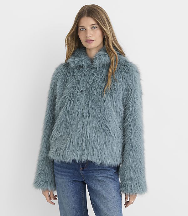 A woman in a teal faux fur jacket and blue jeans stands against a plain white background.