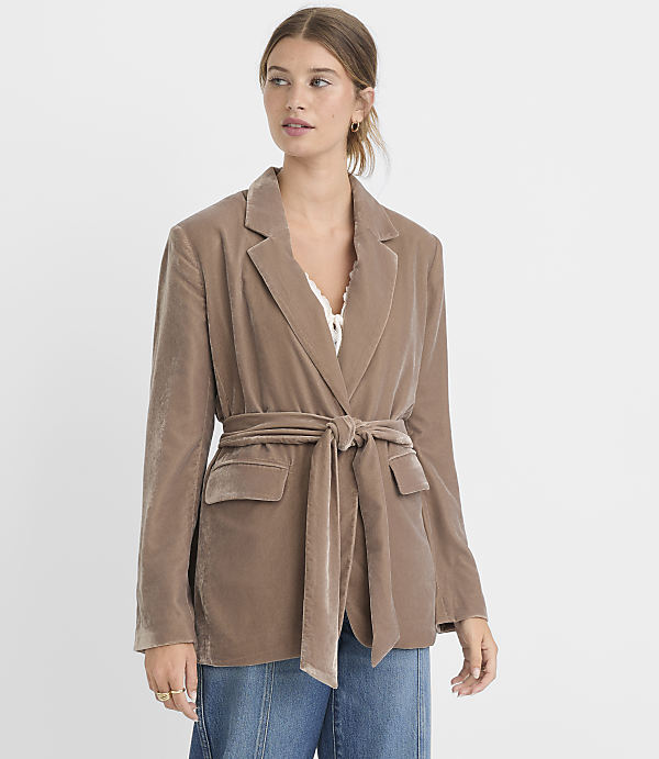 A woman in a taupe velvet blazer cinched with a matching belt stands against a plain white background.