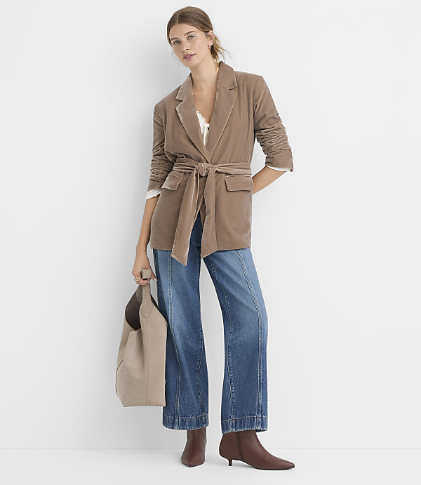 A woman in a brown belted blazer and wide-leg jeans stands against a plain white background, holding a taupe tote bag.