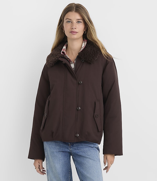 A woman in a brown jacket and jeans stands against a plain white background.