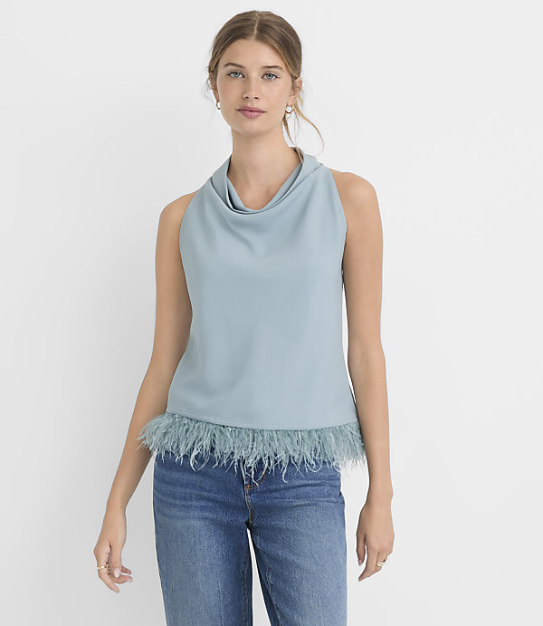 A woman in a blue cowl-neck sleeveless top with a feathered hem and blue jeans stands against a plain white background.
