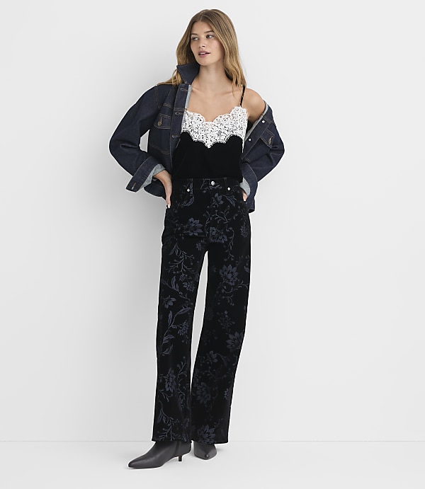 A woman in a denim jacket, lace-trimmed camisole, and floral velvet pants stands against a plain white background.