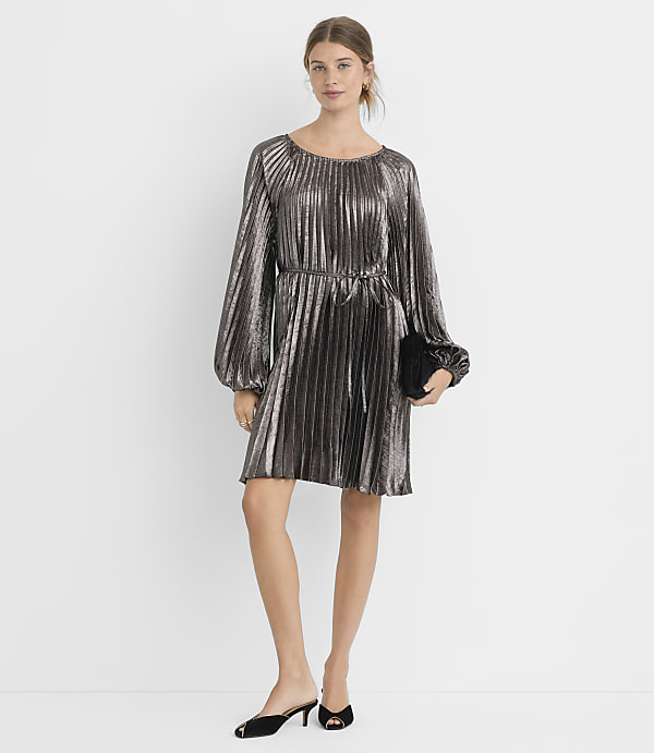 A woman in a silver pleated dress and black mules stands against a plain white background, holding a black clutch.