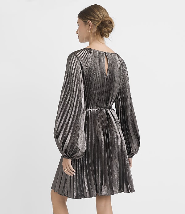A woman in a silver pleated dress stands against a plain gray background.