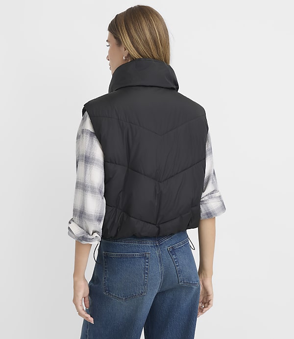 A woman in a black puffer vest and plaid shirt stands against a plain white background.