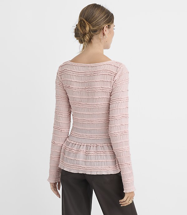 A woman in a pink textured top and dark pants stands against a plain gray background.