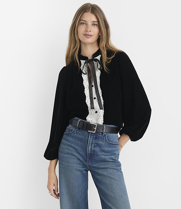 A woman in a black velvet blouse and blue jeans stands against a plain white background.