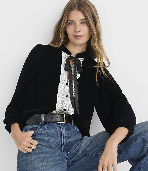 A woman in a black velvet blouse with a lace inset and a black ribbon tie poses in front of a plain white background.