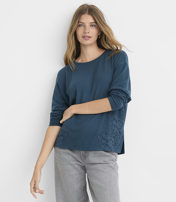 A woman in a teal long-sleeved shirt with lace detailing on the sides stands against a plain gray background.