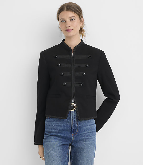 A woman in a black military-style jacket and blue jeans stands against a plain white background.
