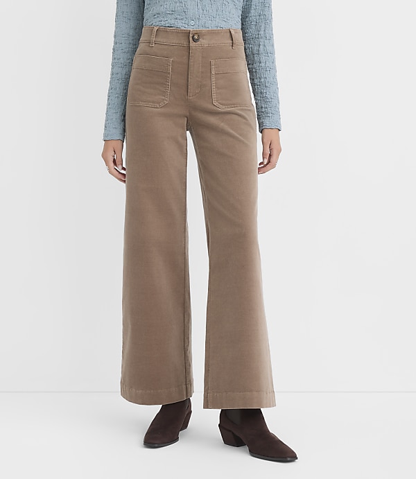 A person stands in front of a white background wearing a blue textured shirt and tan corduroy wide-leg pants with brown boots.