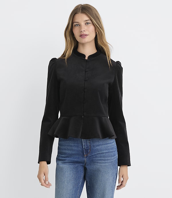 A woman in a black peplum jacket and blue jeans stands against a plain white background.