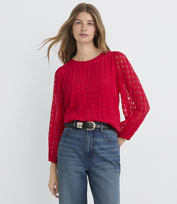 A woman in a red textured blouse and blue jeans stands against a plain white background.