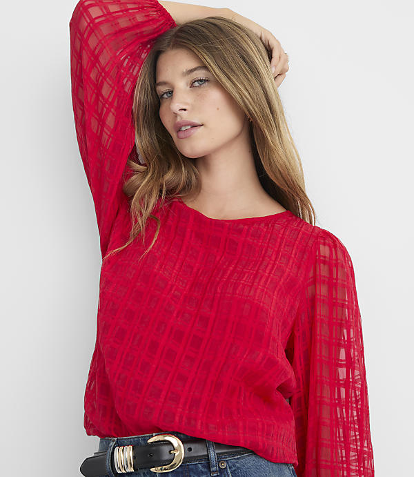 A woman in a red, sheer, plaid blouse with her hand behind her head stands against a plain gray background.
