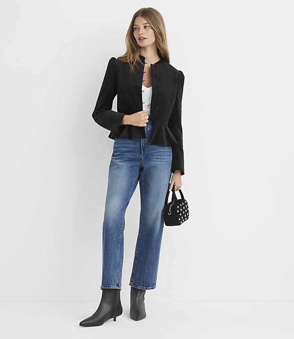 A woman in a black peplum jacket, blue jeans, and black boots stands against a plain white background, holding a small studded handbag.