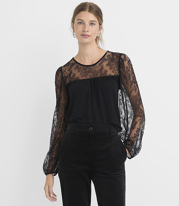 A woman in a black lace-sleeved top and black pants stands against a plain white background.