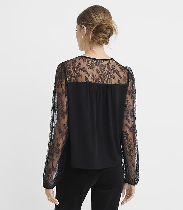 A woman with her hair in a bun wears a black lace-sleeved blouse and black pants against a plain white background.