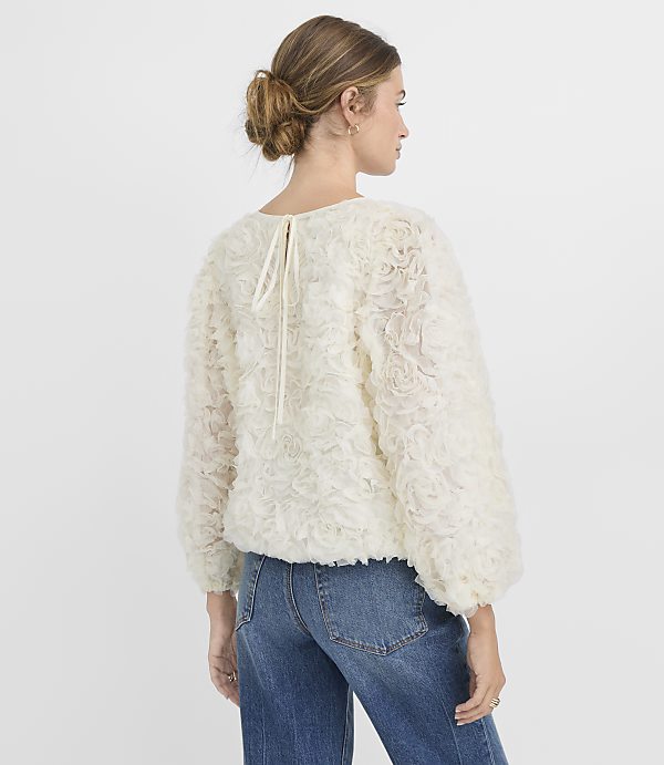 A woman in a textured cream sweater and blue jeans stands against a plain white background.