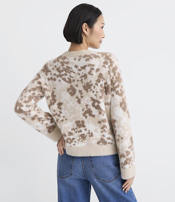 A woman wearing a brown and beige leopard-print fuzzy sweater and blue jeans stands against a plain white background.