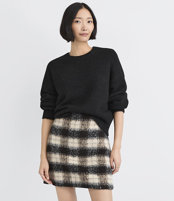 A woman wearing a black sweater and a plaid skirt stands against a plain white background.