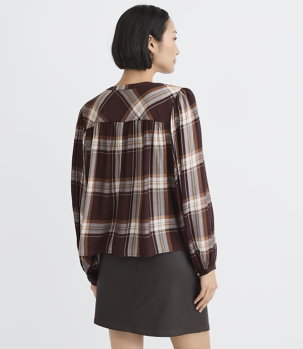A woman wearing a brown plaid blouse and a dark grey skirt stands against a plain white background.
