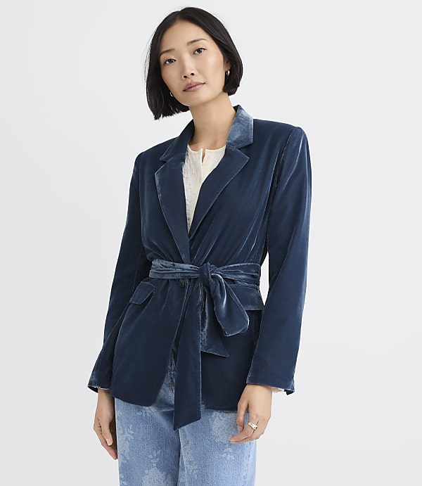 A woman wearing a blue velvet blazer and light-wash jeans stands against a plain white background.