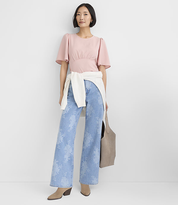 A woman wearing a pink top, floral blue jeans, and a white sweater tied around her waist stands against a plain white background.