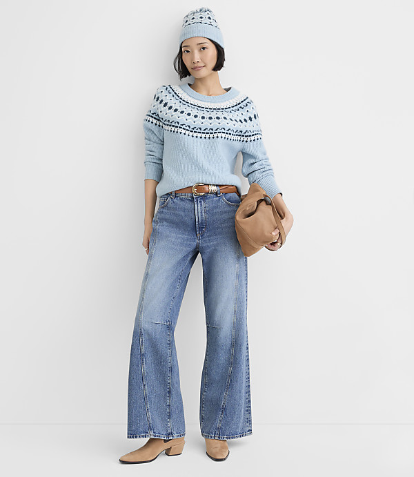 A woman wearing a light blue patterned sweater, a matching beanie, and wide-leg jeans stands against a plain white background.