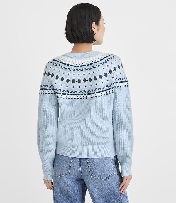 A woman wearing a light blue patterned sweater and blue jeans stands with her back to the camera against a plain white background.