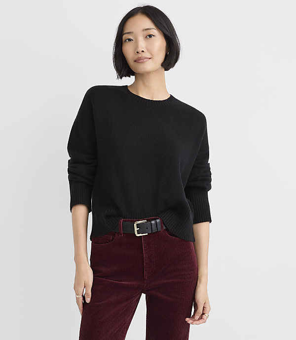 A woman wearing a black sweater and maroon corduroy pants stands against a plain white background.