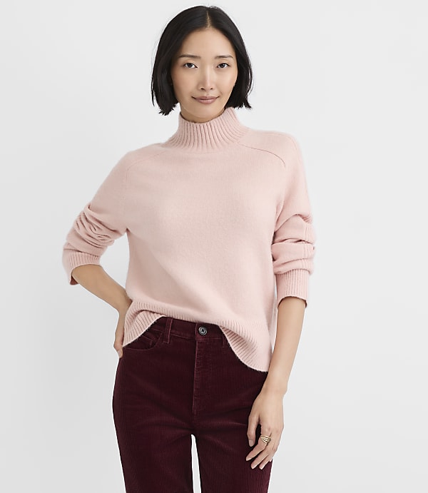 A woman wearing a light pink turtleneck sweater and burgundy corduroy pants stands against a plain white background.